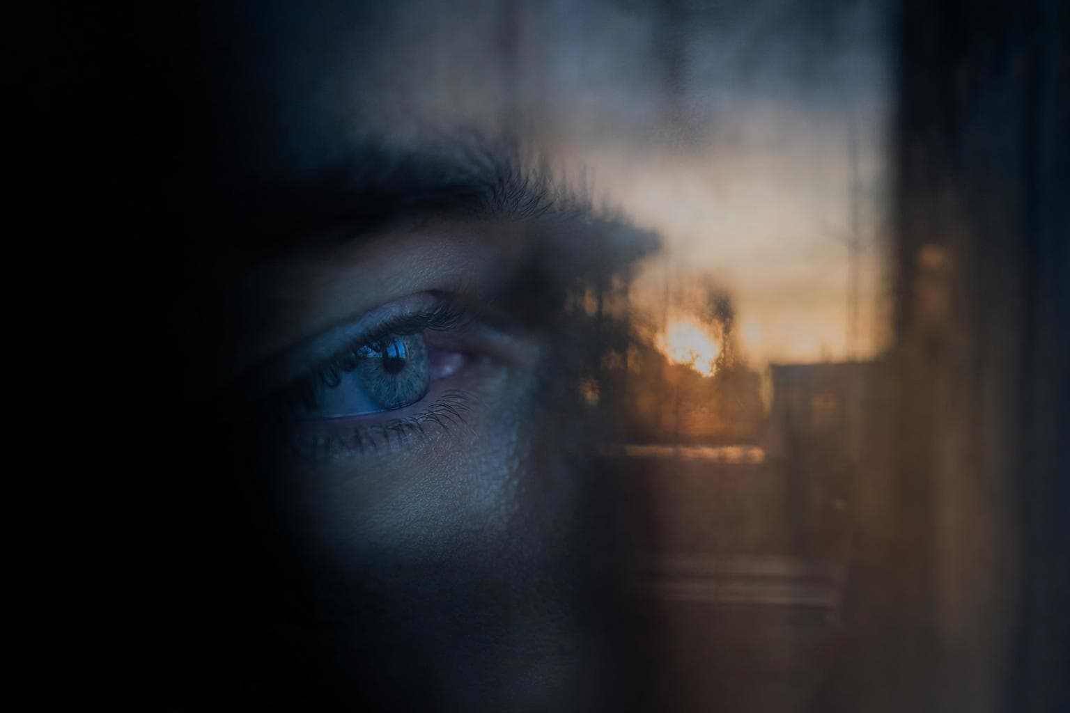 A close-up of a single cool-lit human eye seen through a sheet of clear glass, with a faint warm ghosted reflection of another scene floating across the iris so two images occupy the same surface, neither fully resolving.