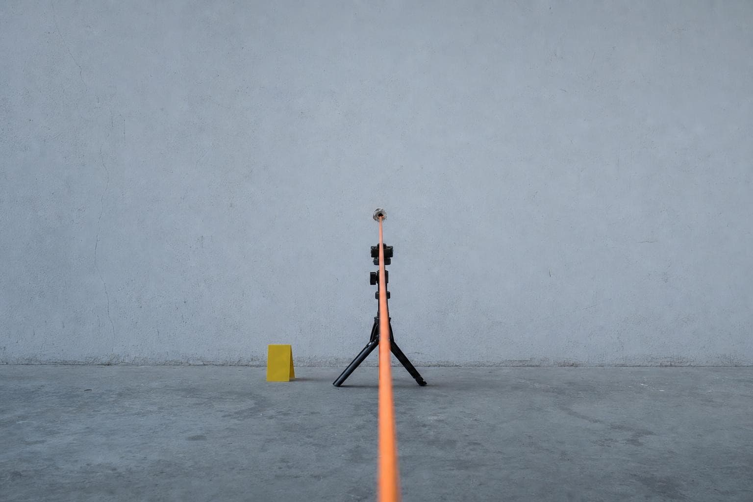 A single slim orange trajectory rod pushed through a bullet hole in a pale plaster wall, held level on a small tripod, casting a long hard shadow across an empty concrete room in flat cold light.
