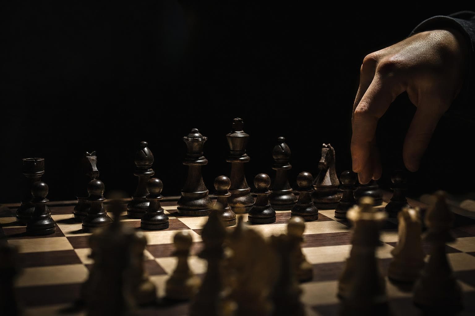 A chess board mid-game shot low from the defending side, the opponent’s pieces advanced and poised, a single hand hovering to answer the threat.
