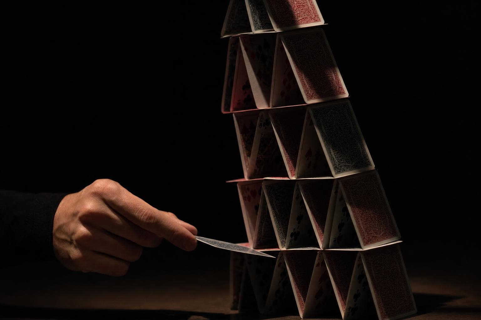 A tall, precarious house of cards under hard light, a single hand drawing one card out from low in the structure as the tower begins to lean.