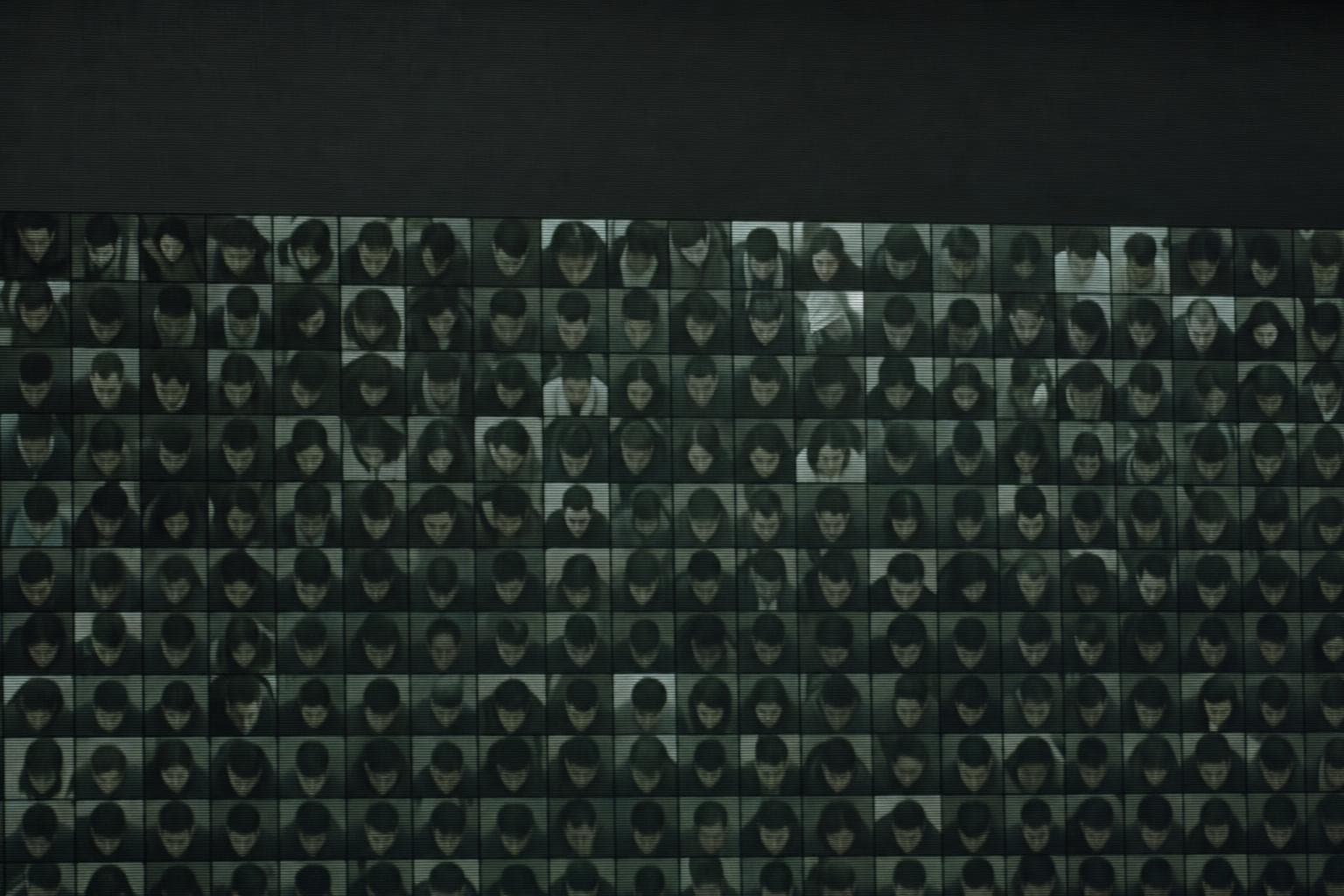 A dense overhead surveillance grid of many near-identical anonymous human faces in desaturated grey-green, each face caught from above in low-resolution CCTV, all of them slightly soft and indistinct, the whole wall reading as one watched, interchangeable crowd.