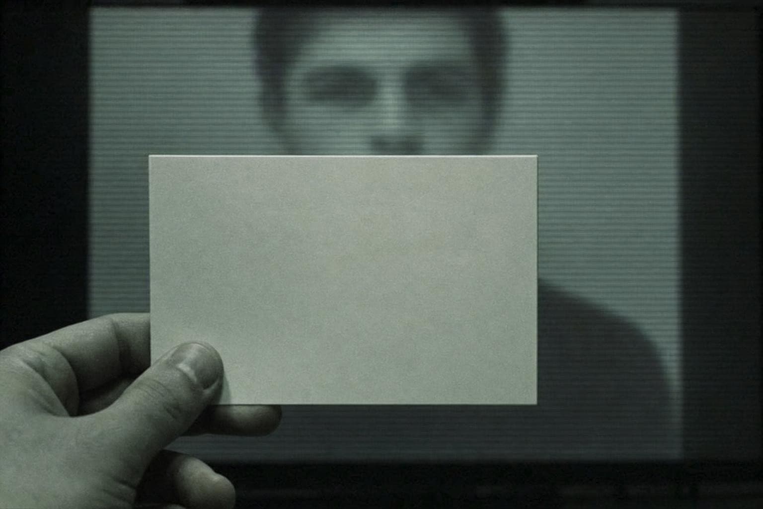 A surveillance screen showing an anonymous face, with a plain opaque card held up in the foreground directly in front of the screen so it reaches the eye first, the card sharply lit and read before the dim face waiting behind its edge.