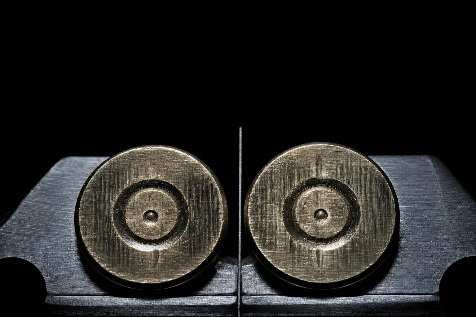 Two brass cartridge case bases side by side under a comparison microscope in cold raking light, their breech-face marks and firing-pin impressions catching the light, a fine dividing line down the middle of the split field.