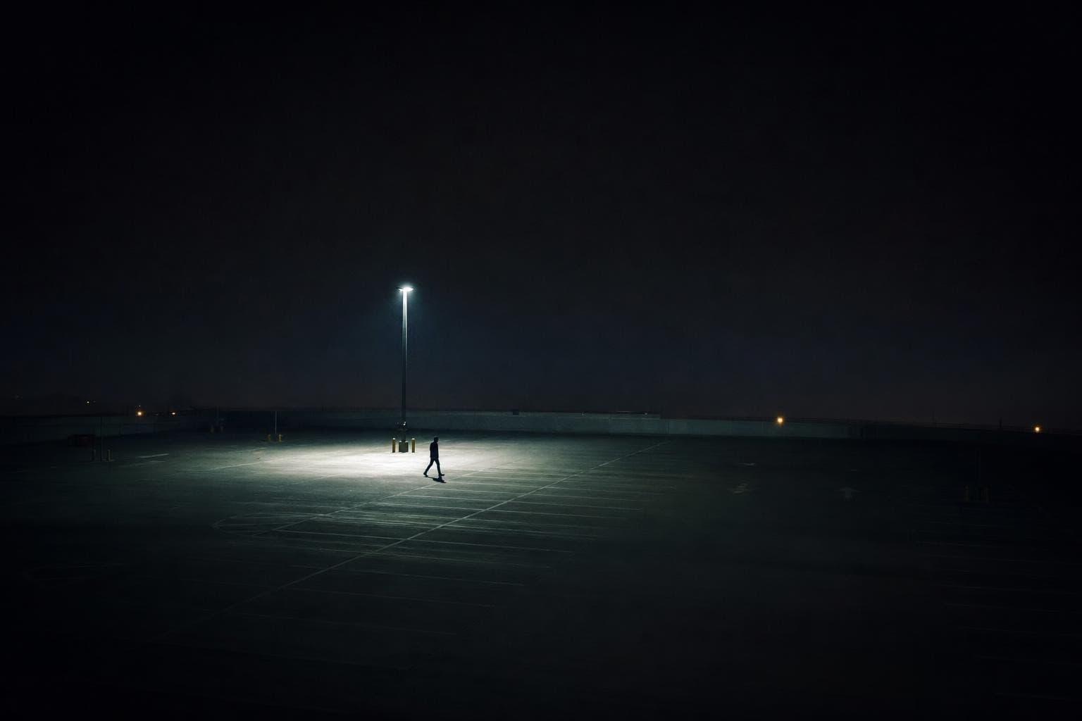 A lone anonymous figure walks across a vast empty car park at midnight, small in deep darkness, crossing through the single pale pool of light thrown by one overhead lamp, the rest of the lot lost in cold blue-black.