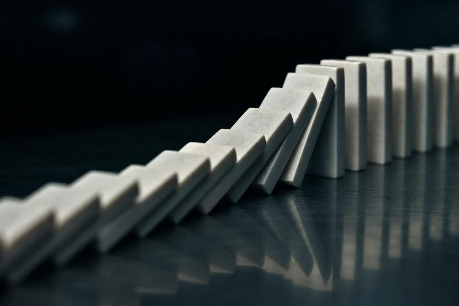 A long line of identical dominoes caught mid-fall, each toppling into the next in an unbroken chain running away across a dark surface.